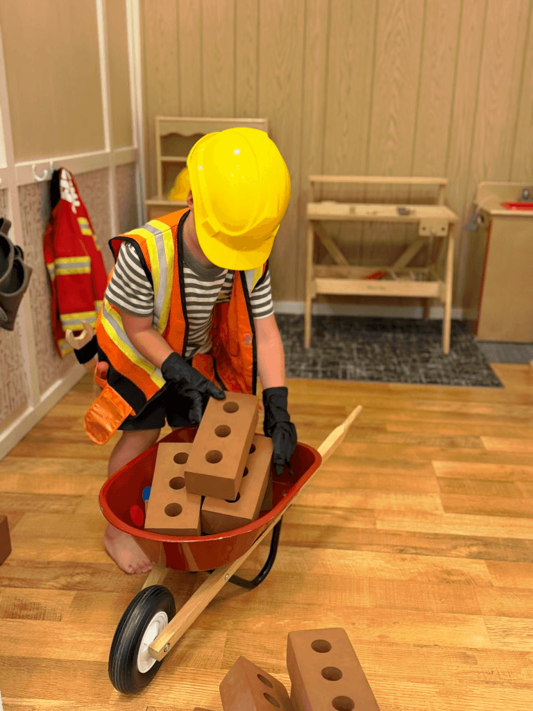 Child in a construction costume playing with a toy wheelbarrow and bricks